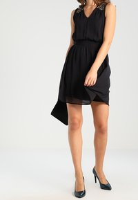 Mustapuku, jossa on V-mallinen kaula-aukko ja koristellut olkapäät. Valoisa siluetti, jossa on korkea-matala helma, yhdistettynä teal-värisiin korkokenkiin.