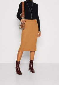 Un pull noir en maille, une jupe midi côtelée beige, et des bottines à talons bordeaux. La tenue est accompagnée d'un sac en cuir marron texturé et d'un collier en argent.