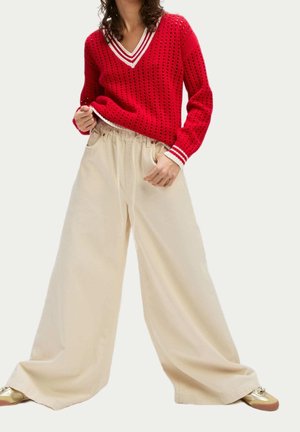 Kvinde iført rød strikket V-hals sweater med hvide striber og brede beige bukser, stående med hænderne nær lommerne.