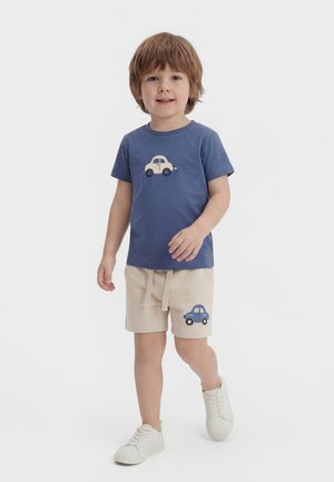 Młody chłopiec idzie, ubrany w niebieską koszulkę i beżowe szorty, obie z grafikami samochodów, oraz białe trampki na jednolitym białym tle.