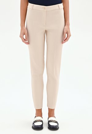 Donna che indossa pantaloni beige aderenti con chiusura a bottone singolo, mocassini bianchi e neri, e una maglia blu navy, in piedi contro uno sfondo bianco.