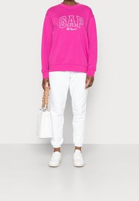 Persona che indossa una felpa GAP rosa acceso, pantaloni jogger bianchi, sneakers bianche, con una borsa bianca a tracolla con manico intrecciato color terra, in piedi su un pavimento grigio.