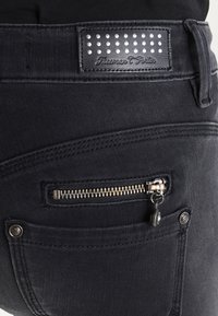 Svarta denimjeans har en liten ficka med dragkedja, kontrasterande sömmar och en läderetikett med nitar vid midjan.