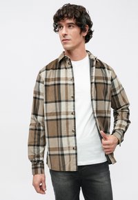 Καφέ και μπεζ καρό πουκάμισο με κουμπιά, με υφή υφάσματος, που διαθέτει γιακά και μακριά μανίκια, φορεμένο πάνω από ένα μονόχρωμο λευκό t-shirt.