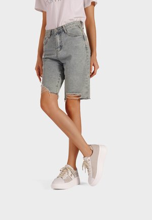Bermuda shorts in denim bluastro con orli sfrangiati, abbinati a sneakers beige con un pannello di accento testurizzato e suola bianca.