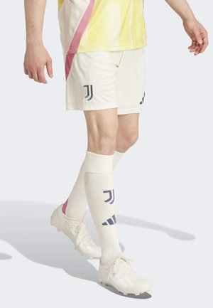 Weiße Fußballshorts mit dunklem blauen Logo, akzentuiert mit Pink, kombiniert mit weißen Socken, die Logos und einen pinken Streifen aufweisen, getragen mit weißen Fußballschuhen.