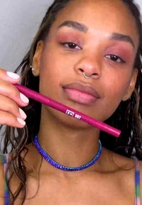 Stylos eyeliner couleur baie tenu dans la main, avec des accents argentés. Un collier multicolore et un maquillage naturel sont également visibles.