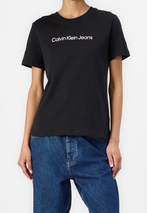 Person wearing black short-sleeve t-shirt with "Calvin Klein Jeans" text and blue denim jeans, standing against a plain background.