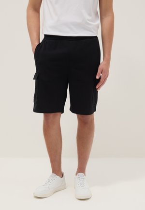 Männliche Person trägt schwarze Cargoshorts mit Seitentaschen, ein weißes T-Shirt und weiße Turnschuhe, steht vor einem schlichten hellen Hintergrund.