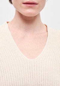 Μπεζ πλεκτό πουλόβερ με σχέδιο v-neck, διαθέτοντας υφή υφάσματος και χαλαρή εφαρμογή, που αναδεικνύει τα σχέδια ραφής γύρω από την κοπή λαιμού.