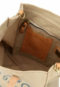 Beige Leinwand-Tragetasche mit einem Reißverschlussfach aus hellbraunem Leder und einem Aufnäher. Verfügt über Ziernähte an den Kanten und einen Messingring als Beschlag.