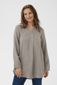 Leende kvinna med vågigt brunt hår bär en lös, långärmad grå blus och blåjeans, stående mot en enkel ljusgrå bakgrund.