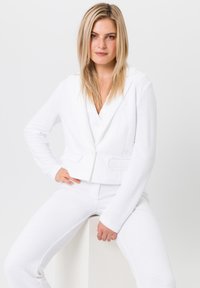 Weißer strukturierter Anzug mit einem verkürzten Blazer, der einen geschlitzten Kragen und zwei Vorderentaschen aufweist, kombiniert mit passenden, taillierten Hosen.