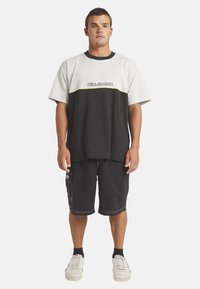 Musta-valkoinen lyhythihainen paita, jossa on keltainen korostus ja "BILLABONG" -logo, yhdistettynä mustiin shortseihin, joissa on sivutaskut ja valkoinen ommel.