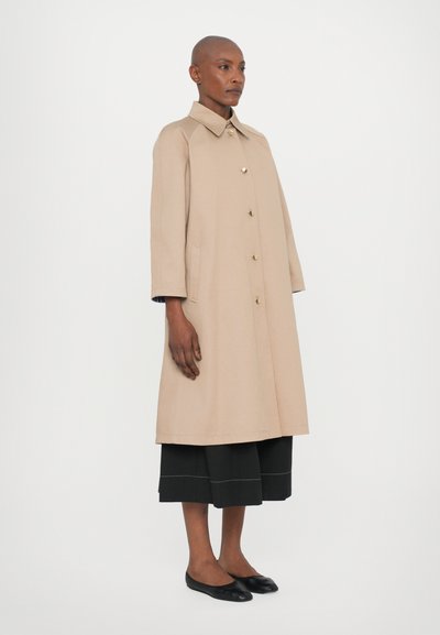 Cappotto trench beige con colletto, bottoni dorati e tasche laterali. Accompagnato da culotte nere e scarpe flat, su uno sfondo neutro.