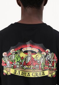 Schwarzes T-Shirt mit einem bunten Grafikmotiv von Skeletten an einem Tisch, mit Details wie Blumen, Getränken und dem Text "SANTA CRUZ."