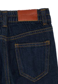 Jean en denim bleu foncé avec une poche arrière, des coutures orange et une étiquette en cuir marron embossée de "TRUSSARDI" à la taille.
