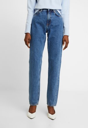 Persona con jeans azules de talle alto, suéter azul claro y tacones blancos de punta afilada, sobre un fondo blanco liso.