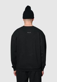 Svart sweatshirt med avslappnad passform, ribbade muddar och fåll, med texten "F.U.T." tryckt i vitt på baksidan. Bärs med en svart mössa.