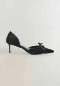 Czarne, aksamitne szpilki z ostro zakończonym noskiem i detalem w postaci kokardy na jasnym tle.