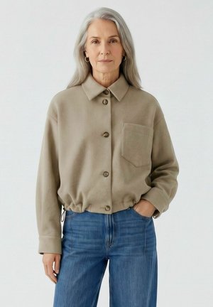Ältere Frau mit langen grauen Haaren, trägt eine beige geknöpfte Jacke und blaue Jeans, steht mit einer Hand in der Tasche vor einfachem Hintergrund.