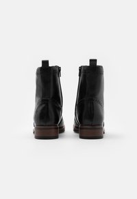 Un par de botas negras de cuero hasta el tobillo con tacones apilados marrones, perforaciones decorativas y lengüetas para tirar, fotografiadas desde la parte posterior sobre un fondo blanco.