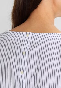 Blouse rayée blanche avec col bateau, fermeture au dos par boutons de couleur claire, et texture de tissu lisse.