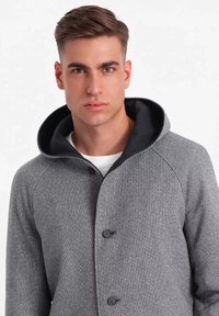 Chaqueta gris texturizada con capucha negra y botones. Cuenta con un corte moderno y mangas raglán, ofreciendo un aspecto en capas.