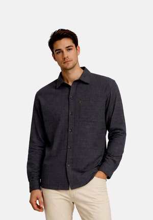 Young man wearing a dark gray checkered long-sleeve button-up shirt with beige pants, one hand in pocket, standing against plain background.