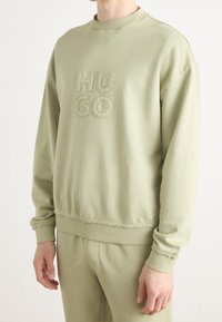 Persoon draagt een lichtgroene trui en bijpassende broek, trui geborduurd met "HUGO" op de borst.