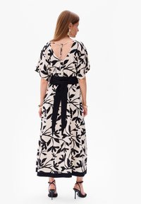 Maxi-jurk van lichte stof met zwart bladpatroon, V-back, brede mouwen en een zwarte ceintuur in de taille. Sleehakken maken de look compleet.