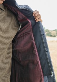 Chaqueta de lana gris con cremallera en la parte delantera. El interior cuenta con un forro de patrón burdeos y una sección negra, con un bolsillo interior visible.