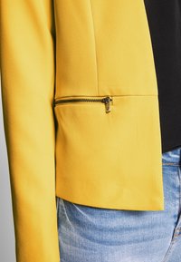 Blazer jaune au design structuré, avec une poche zippée et une texture lisse, associé à un denim bleu.