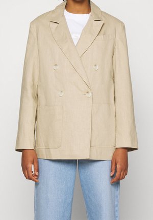 Person wearing a beige double-breasted blazer with four buttons, large front pockets, white shirt, and light blue jeans.