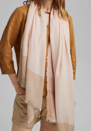 Femme portant une écharpe rose clair à bords frangés par-dessus un haut à manches longues moutarde et un short beige, debout avec une main dans la poche.