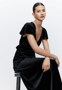 Vestito in velluto nero con maniche corte arricciate, scollo a V profondo e vita aderente che si allarga in una gonna fluente. Texture liscia.