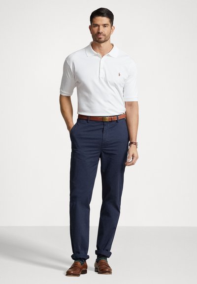 Man staat met handen in de zakken, draagt een witte kortemouwen polo, donkerblauwe broek, bruine riem, bruine schoenen en een horloge tegen een effen achtergrond.