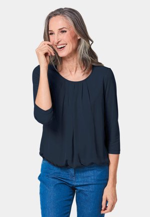 Marineblauwe blouse met driekwartmouwen, ronde halslijn en geplooide voorkant. Draag met blauwe jeans, gemaakt van een lichte stof.
