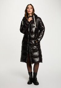 Abrigo tipo puffer negro y brillante con capucha, que cuenta con una cremallera frontal, dos bolsillos laterales y cordones de marca en blanco. Diseñado con una silueta larga y volumétrica.