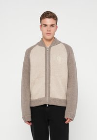 Jovem vestido com um suéter de malha com fecho de correr, com painéis frontais bege, mangas e colarinho taupe, combinado com calças pretas, diante de um fundo branco.