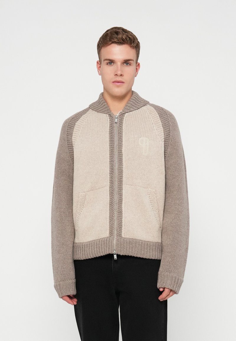Jovem vestido com um suéter de malha com fecho de correr, com painéis frontais bege, mangas e colarinho taupe, combinado com calças pretas, diante de um fundo branco.