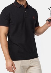 Černé polo tričko s červeným zdobným švem, krátké rukávy, tři knoflíky. Nosené s světlými kalhotami. Ruka držící sluneční brýle.