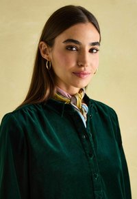 Grüne Samtbluse mit Kragen, Knopfleiste und gerafftem Schulterdetails. Ein verknoteter, mehrfarbiger Schal sorgt für Kontrast.