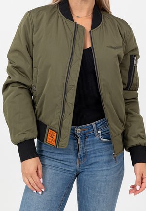 Kvinde iført en olivengrøn bomberjakke med sort ribbet krave og manchetter, kombineret med blå jeans og en sort top under.