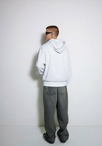 Szary sweatshirt z kapturem i białym pasem, zestawiony z luźnymi ciemnymi dżinsami i czarnymi okularami przeciwsłonecznymi, widoczny z tyłu na tle gładkiej ściany.
