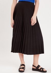 Jupe midi plissée noire avec une texture lisse et une large taille, accompagnée de sandales noires à lanières avec un talon compensé bas.
