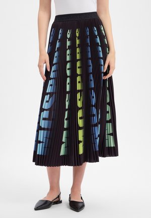 Femme portant une jupe midi plissée noire avec un grand texte vertical bleu et vert, un haut blanc et des chaussures plates slingback noires.