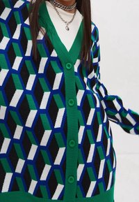 Person klädd i en grön, blå, svart och vit geometriskt mönstrad kofta med gröna knappar och lager av silverkedjor.