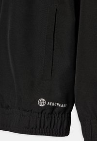 Chaqueta negra con textura suave, que presenta un bolsillo lateral, cinturilla elástica y el logo blanco "AEROREADY" con un diseño de globo terráqueo.