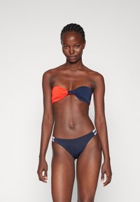 Top bikini senza spalline in color block arancione e blu navy, con nodo al centro. Parte inferiore in blu navy solido con fascia logo a contrasto. Texture liscia.